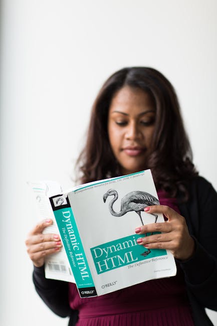 The image shows a woman with dark brown, slightly wavy hair, wearing a black blazer over a dark pink top, holding an open technical book titled 'Dynamic HTML' published by O'Reilly. She is in a well-l